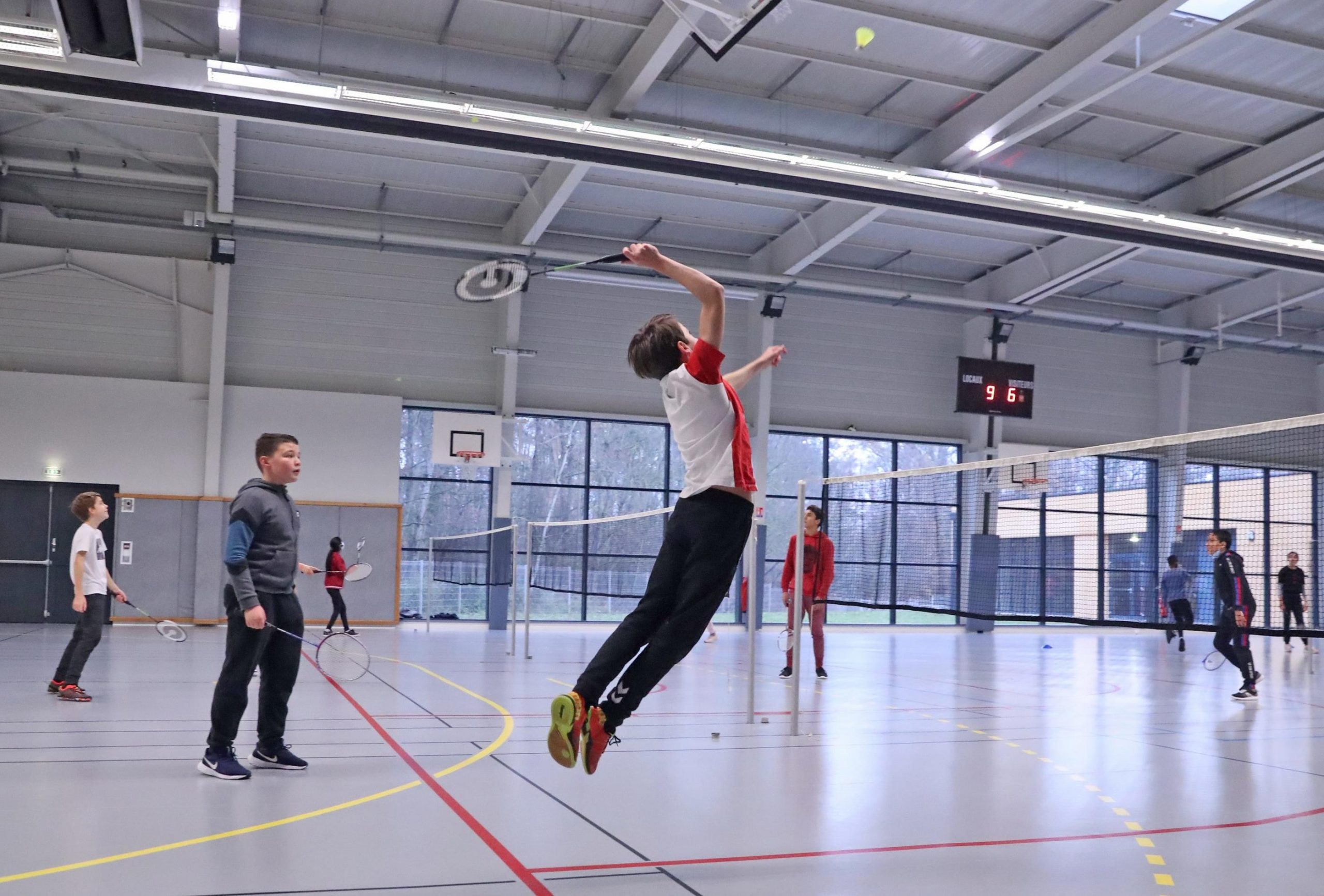 Photo illustrant un jeune ado qui saute pour frapper un volant à l'aide de sa raquette de badminton. Il se trouve dans un gymnase avec plusieurs camarades de classe en arrière plan qui jouent également au badminton