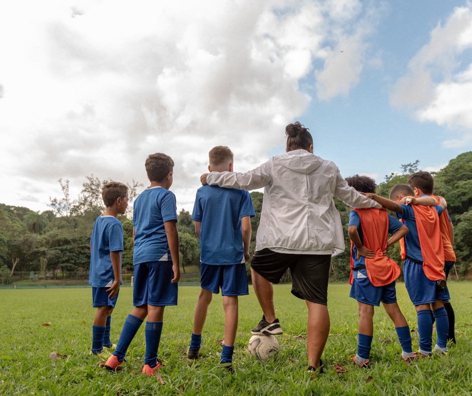 Une coach de foot avec 5 enfants qui est en train de leur faire un brief d'avant match. Les personnes sont vues de dos.